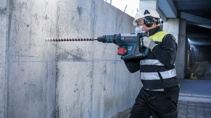 A person wearing safety equipment drills into a concrete wall with a rotary hammer.
