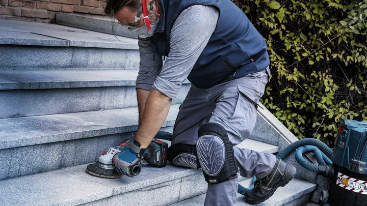 A worker wearing safety equipment grinds outdoor stone stairs with a power tool.
