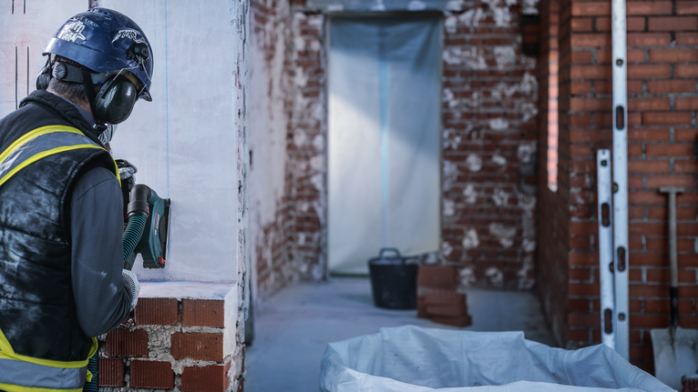 Person wearing safety equipment drills into a wall at a construction site.