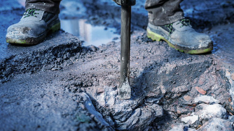 A person wearing safety equipment breaks concrete with a demolition tool.