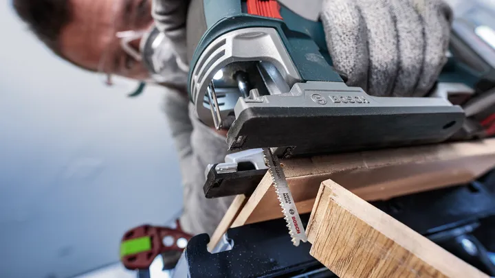 Person wearing safety equipment uses a power jigsaw to cut a piece of wood.