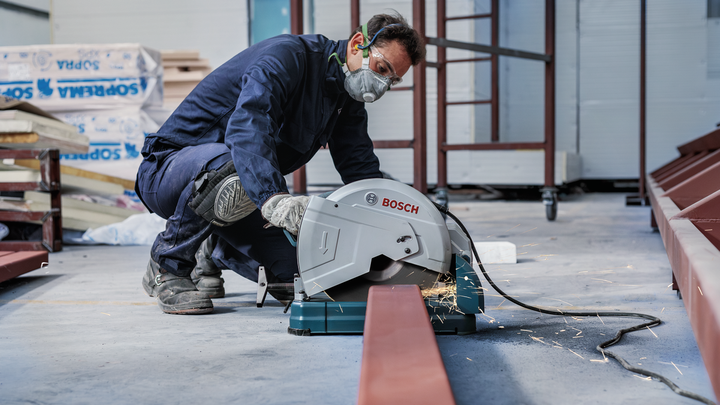 A person wearing safety equipment cuts a steel beam with a circular saw.