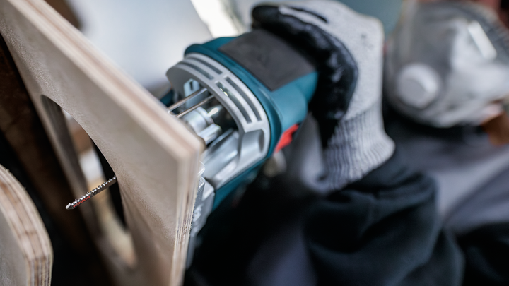 A person wearing safety equipment cuts plywood with a jigsaw.