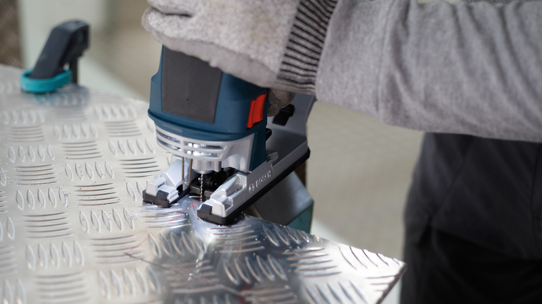Person wearing safety equipment uses a jigsaw to cut patterned metal sheet.