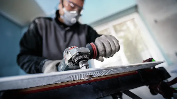 Person wearing safety equipment grinds the edge of a large tile on a workbench.