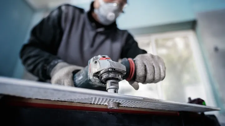 Person wearing safety equipment grinds the edge of a tile with a power tool.