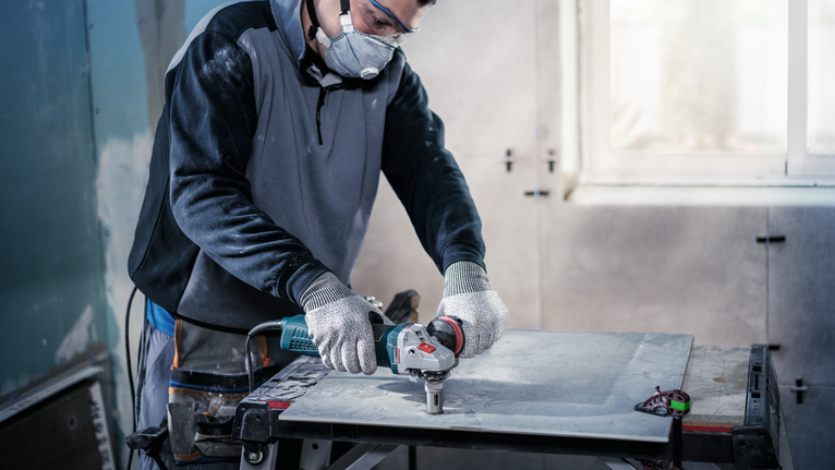 Person wearing safety equipment cuts a tile with an angle grinder indoors.