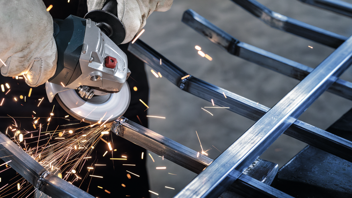 Person wearing safety equipment uses an angle grinder to cut metal, producing sparks.