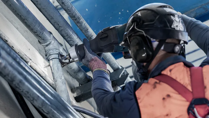 A person wearing safety equipment cuts a metal pipe with a reciprocating saw.