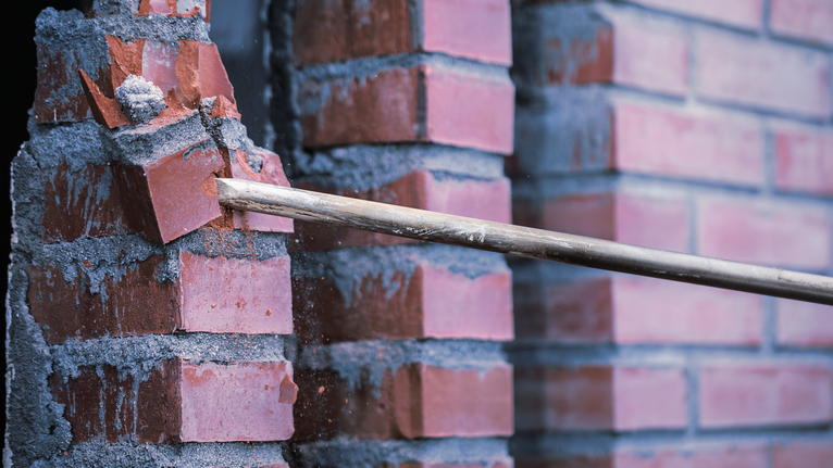 Demolition bar breaking red bricks from a mortar wall.
