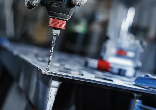 Person wearing safety equipment drills a hole in a metal sheet.