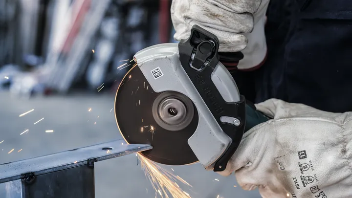 Person wearing safety equipment cuts metal with an angle grinder, producing sparks.