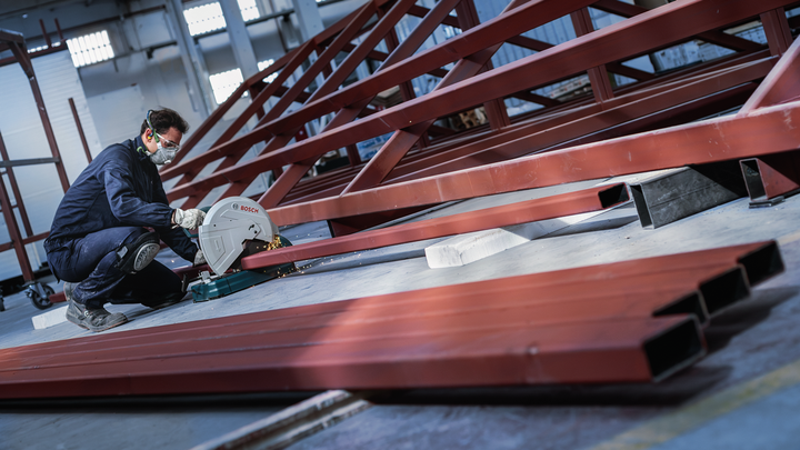 A person wearing safety equipment cuts red metal beams with a circular saw.