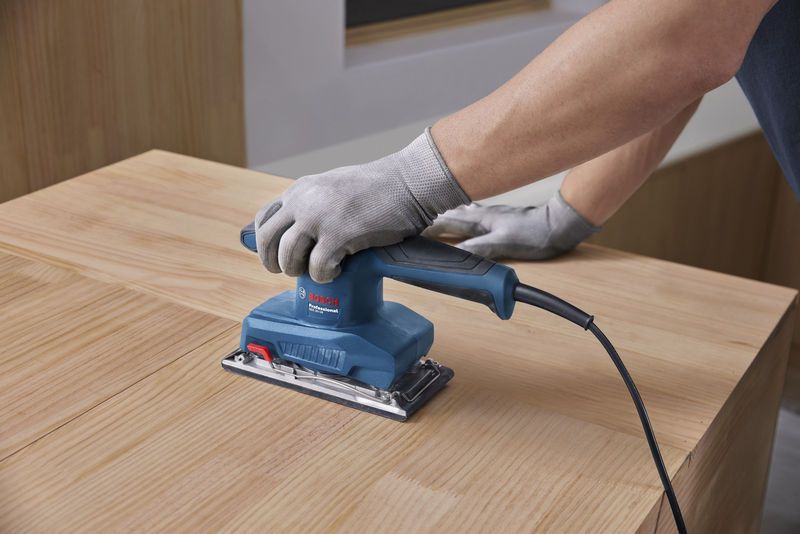 A person wearing safety equipment uses an orbital sander on a wooden surface.
