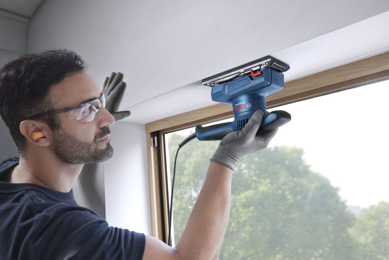 A person wearing safety equipment sands a window frame with an orbital sander.