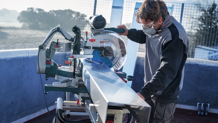 A person wearing safety equipment operates a circular saw to cut a long metal beam.