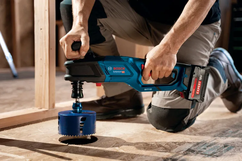 A person wearing safety equipment drills a large hole in plywood with a cordless angle drill.