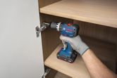 A person wearing safety equipment uses a cordless impact drill to install a cabinet hinge.
