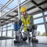 A person wearing safety equipment operates a magnetic core drill on a steel beam.