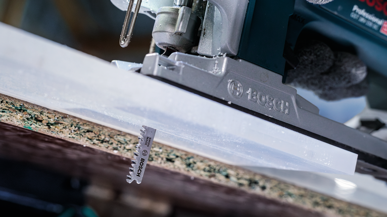 Person wearing safety equipment operates a jigsaw cutting through a laminated board.