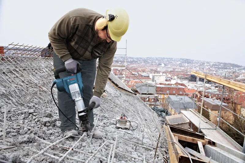 Una persona che indossa un equipaggiamento di sicurezza utilizza un martello demolitore per rompere il cemento su un tetto.