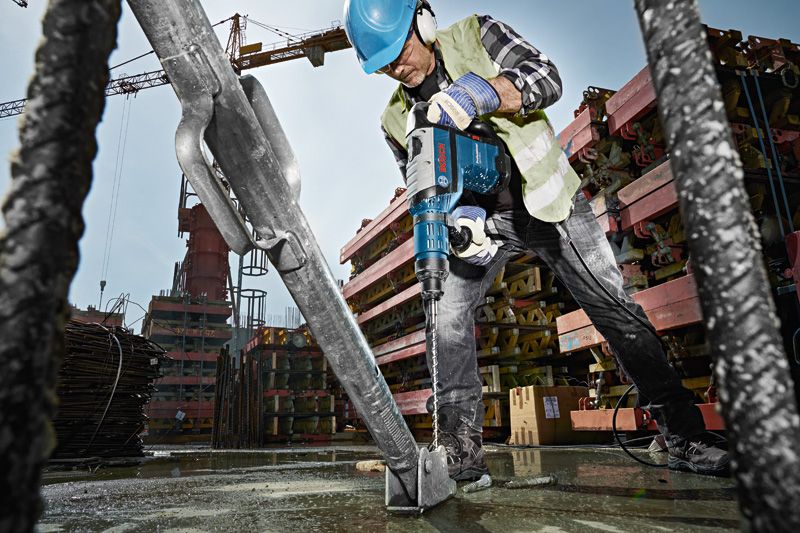Un operaio che indossa dispositivi di sicurezza utilizza un martello perforatore per perforare una base di cemento in un cantiere edile.
