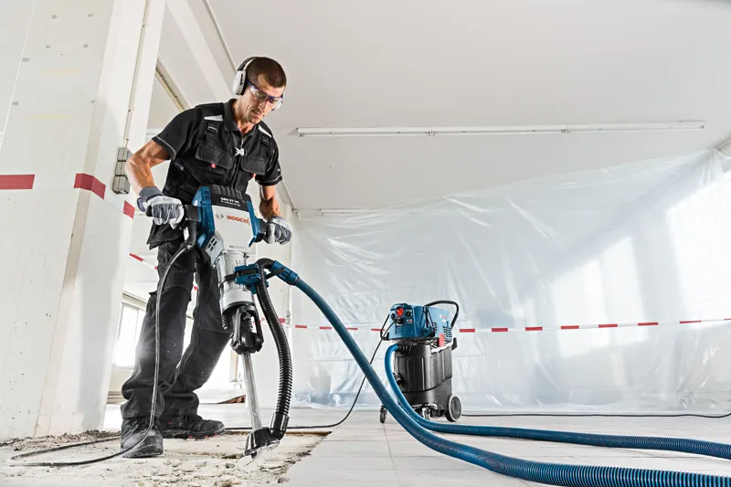 Una persona che indossa un equipaggiamento di sicurezza utilizza un martello demolitore con aspirazione della polvere su un pavimento di cemento.