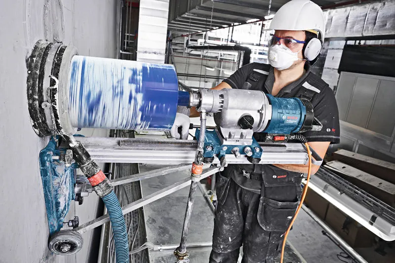 Una persona che indossa un equipaggiamento di sicurezza pratica un grande foro nel cemento utilizzando un trapano diamantato.