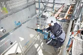 Una persona che indossa un equipaggiamento di sicurezza perfora il cemento con un grosso martello perforatore.