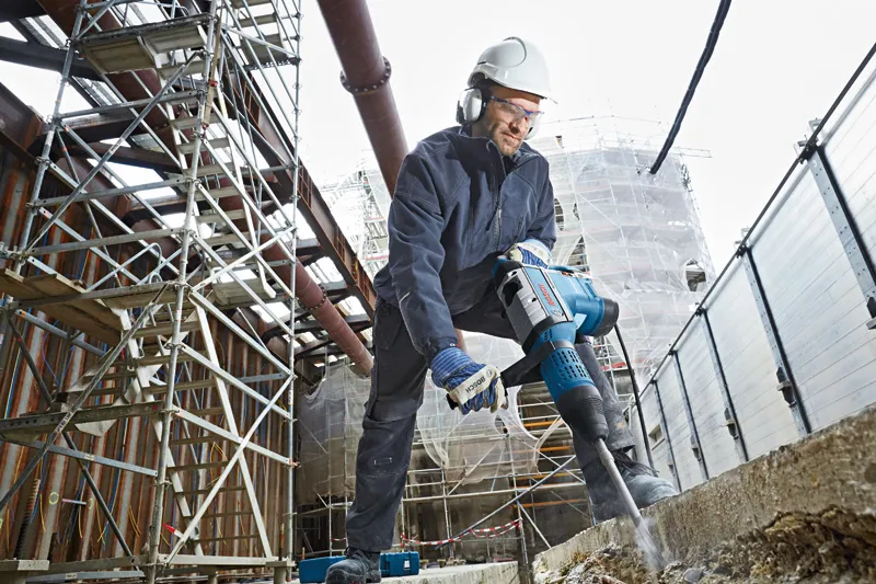 Una persona che indossa un equipaggiamento di sicurezza utilizza un martello perforatore per rompere il cemento in un cantiere edile.