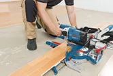 Una persona che indossa dispositivi di sicurezza utilizza una sega troncatrice scorrevole per tagliare il pavimento in laminato.