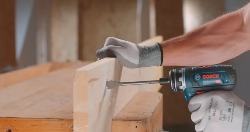 Una persona che indossa un equipaggiamento di sicurezza fora una trave di legno con un utensile elettrico.