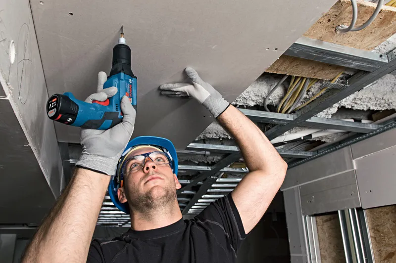 Una persona che indossa dispositivi di sicurezza installa il cartongesso sopra la testa utilizzando un cacciavite a batteria.