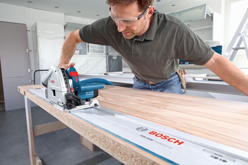 Una persona che indossa un equipaggiamento di sicurezza utilizza una sega a tuffo per tagliare il legno lungo una guida.