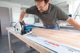Una persona che indossa un equipaggiamento di sicurezza utilizza una sega a tuffo per tagliare il legno lungo una guida.