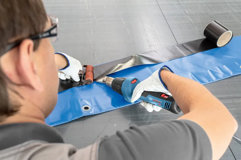 Una persona che indossa un equipaggiamento di sicurezza utilizza una pistola termica per sigillare un foglio di plastica.