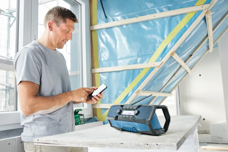 Una persona sistema un telefono vicino a una radio da cantiere blu, appoggiata su un tavolo da costruzione.