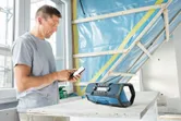 Una persona sistema un telefono vicino a una radio da cantiere blu, appoggiata su un tavolo da costruzione.