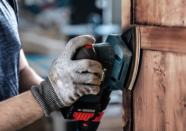 Una persona che indossa dispositivi di sicurezza carteggia una porta di legno con un utensile elettrico.