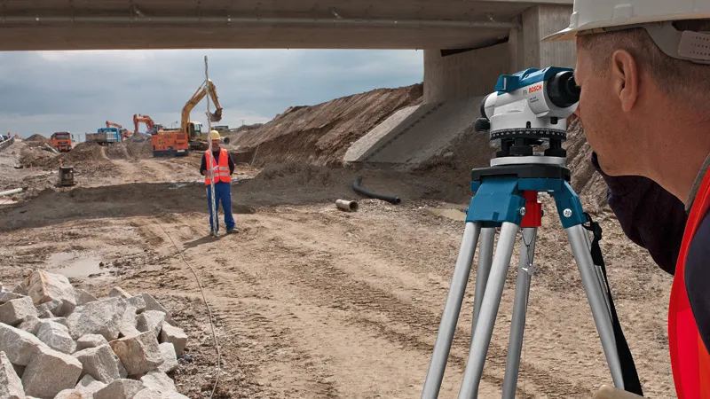Una persona che indossa un equipaggiamento di sicurezza utilizza un livello ottico in un cantiere stradale.