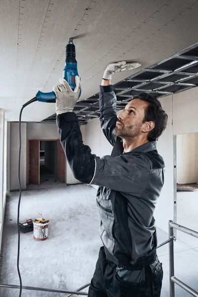 Una persona che indossa dispositivi di sicurezza fissa il cartongesso al soffitto con un cacciavite elettrico.