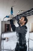 Una persona che indossa dispositivi di sicurezza fissa il cartongesso al soffitto con un cacciavite elettrico.