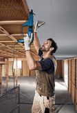 Una persona che indossa dispositivi di sicurezza installa il cartongesso su un soffitto utilizzando un avvitatore elettrico.