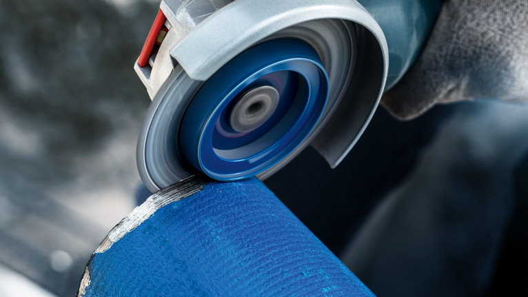 Una persona che indossa l'equipaggiamento di sicurezza leviga un tubo di metallo blu con una smerigliatrice angolare.