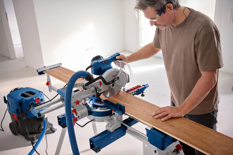 Una persona che indossa dispositivi di sicurezza taglia una tavola di legno con una sega troncatrice.