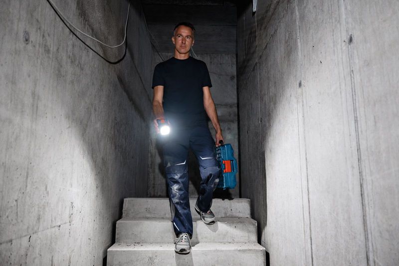 Una persona tiene in mano una lampada da lavoro senza fili e una cassetta degli attrezzi mentre scende le scale di cemento.