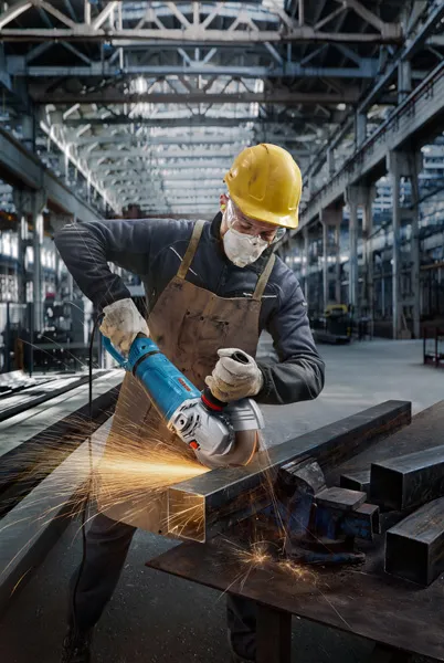 Una persona che indossa un equipaggiamento di sicurezza leviga una trave metallica con una smerigliatrice angolare, creando scintille.