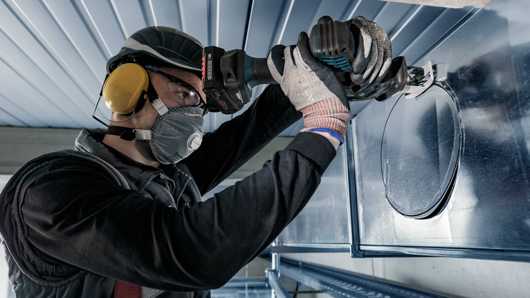 Una persona che indossa un equipaggiamento di sicurezza pratica un foro circolare in una conduttura metallica con un elettroutensile.