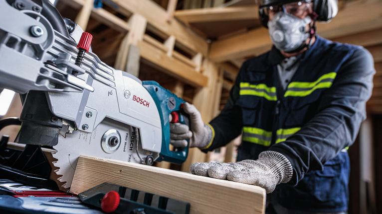 Una persona che indossa un equipaggiamento di sicurezza aziona una sega circolare per tagliare una trave di legno.