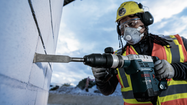 Una persona che indossa un equipaggiamento di sicurezza scalpella un muro di cemento con un utensile elettrico.